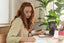 A woman sits at a desk with a laptop and a notebook she is writing in
