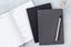 A short stack of three 90 day goal planners, one grey, one black, and one sitting open to a planning page.