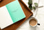 A book sits on a wooden tray next to a cup of tea and a sprig of green. The book is open to a quote about perseverance.