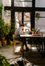 A woman smiles working at a large work table surrounded by lush plants and big windows