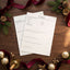 Two blank white sheets of paper stacked on a dark wooden table, surrounded by festive Christmas decorations including red velvet ribbons, pine branches with gold pinecones and berries, and shiny gold ornaments.