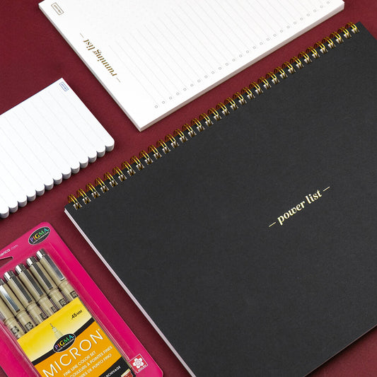 Black spiral-bound notebook with 'power list' on a red surface, next to white notebooks and a pink box of Micron pens.